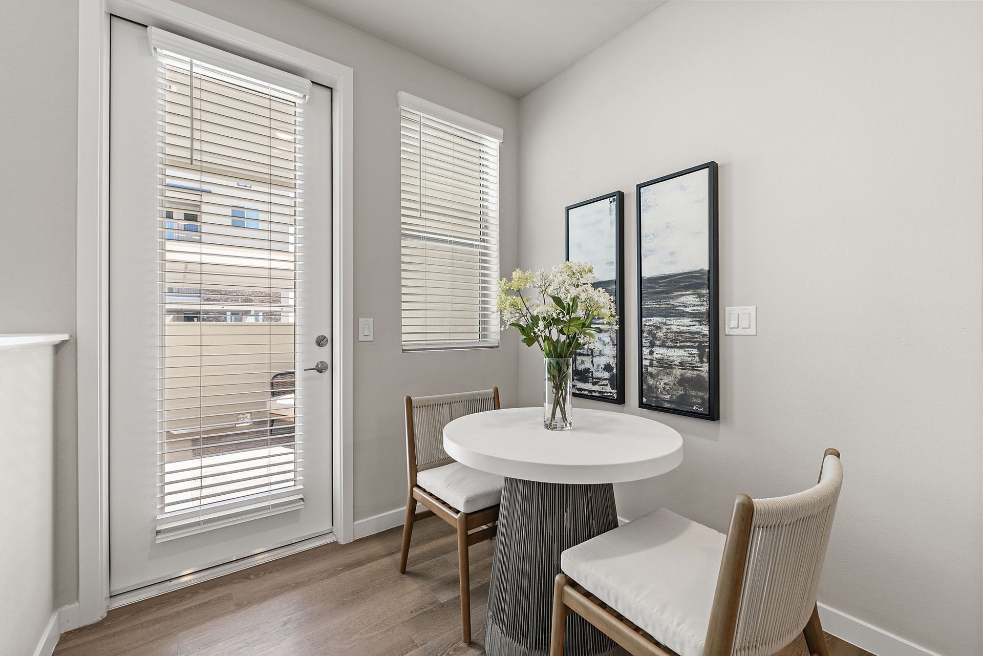 Small dining area with a round white table, two chairs, and a vase of flowers, next to a window and door at Marq Bronco Butte, offers loft-style apartments in North Phoenix, AZ.