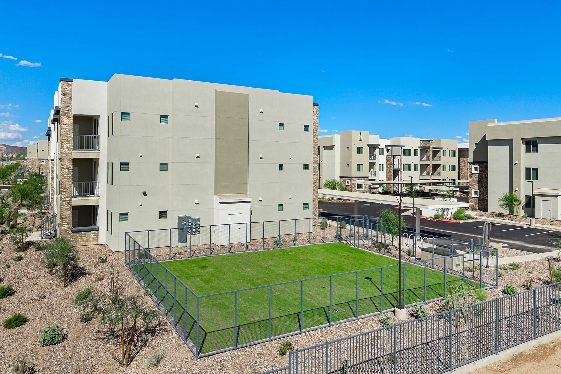 Apartment complex with a small fenced-in grassy area at Marq Bronco Butte, offers 1, 2, and 3 bedroom apartments in North Phoenix, AZ. Tan buildings with balconies, against a bright blue sky.