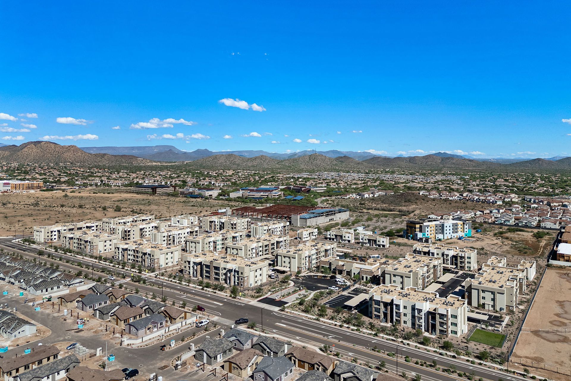 Aerial view of a suburban development with beige buildings, roads, and mountains in the background under a blue sky at Marq Bronco Butte, offers 1, 2, and 3 bedroom apartments in North Phoenix, AZ.
