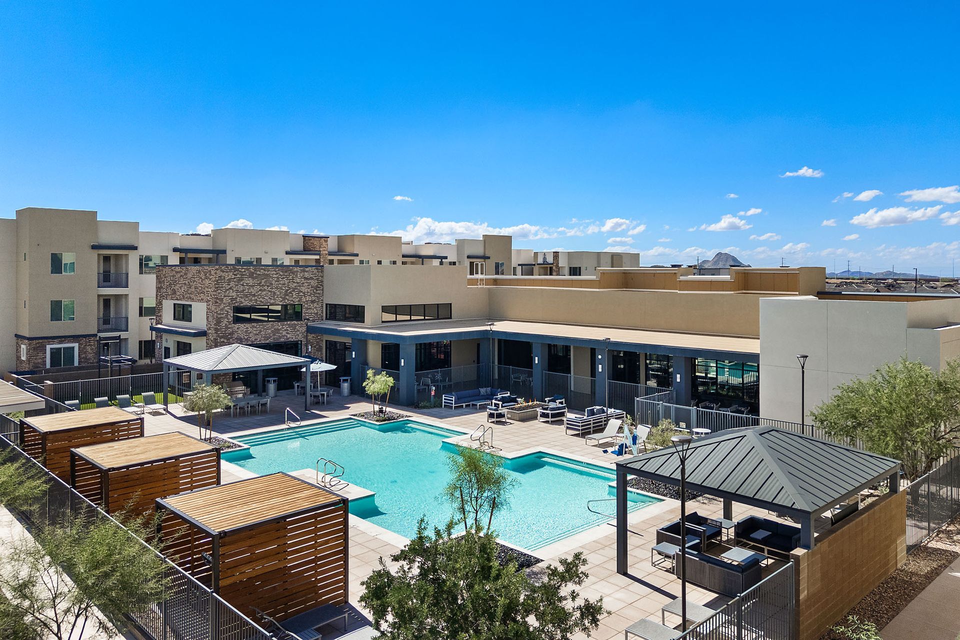 Pool area with cabanas, lounge chairs, and people in front of a multi-story apartment building under a bright blue sky at Marq Bronco Butte, offers brand-new apartments near USAA in North Phoenix, AZ.