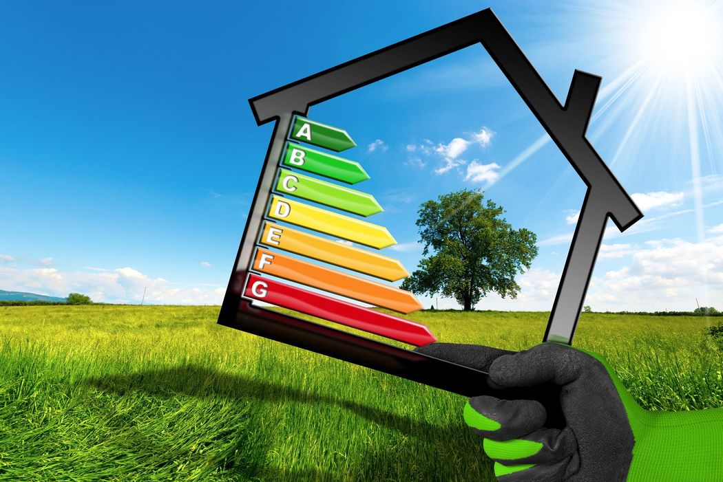 Una mano che regge la struttura di una casa con etichette di efficienza energetica, sullo sfondo di un campo verde e un cielo azzurro.