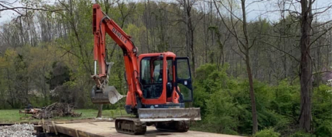 Orange excavator working on a wooden structure near water. Trees and greenery in the background.