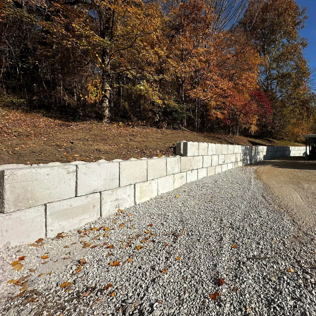 Gray concrete block retaining wall along a gravel driveway, with fall foliage in background.