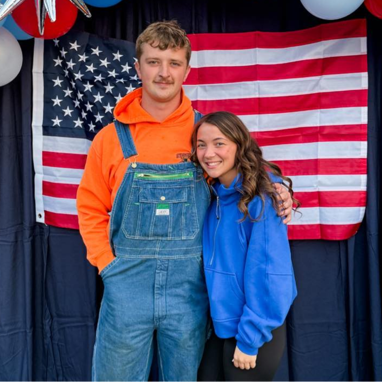 Man in overalls and woman in blue hug in front of an American flag.