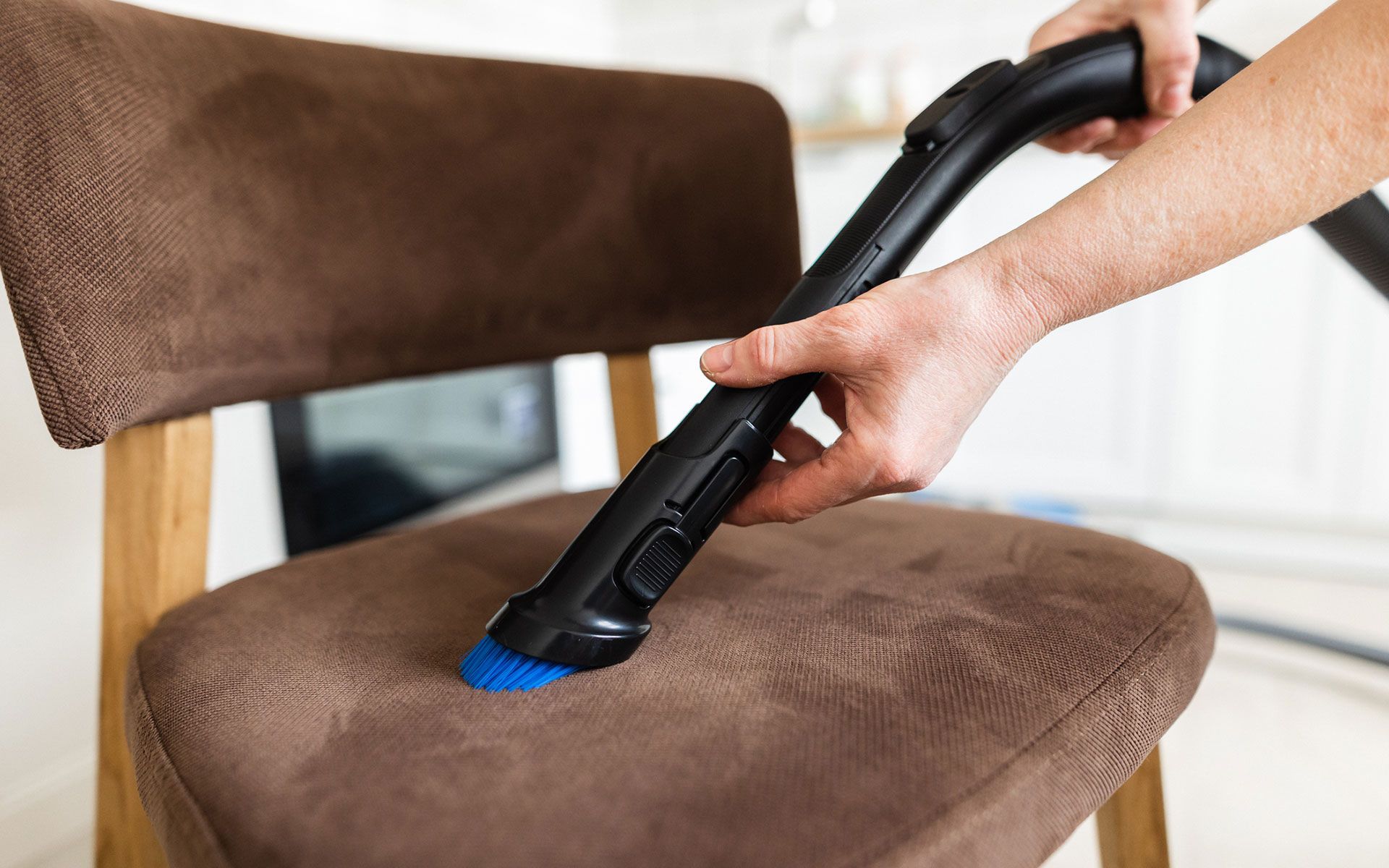Person vacuuming a brown upholstered chair with a black vacuum attachment.