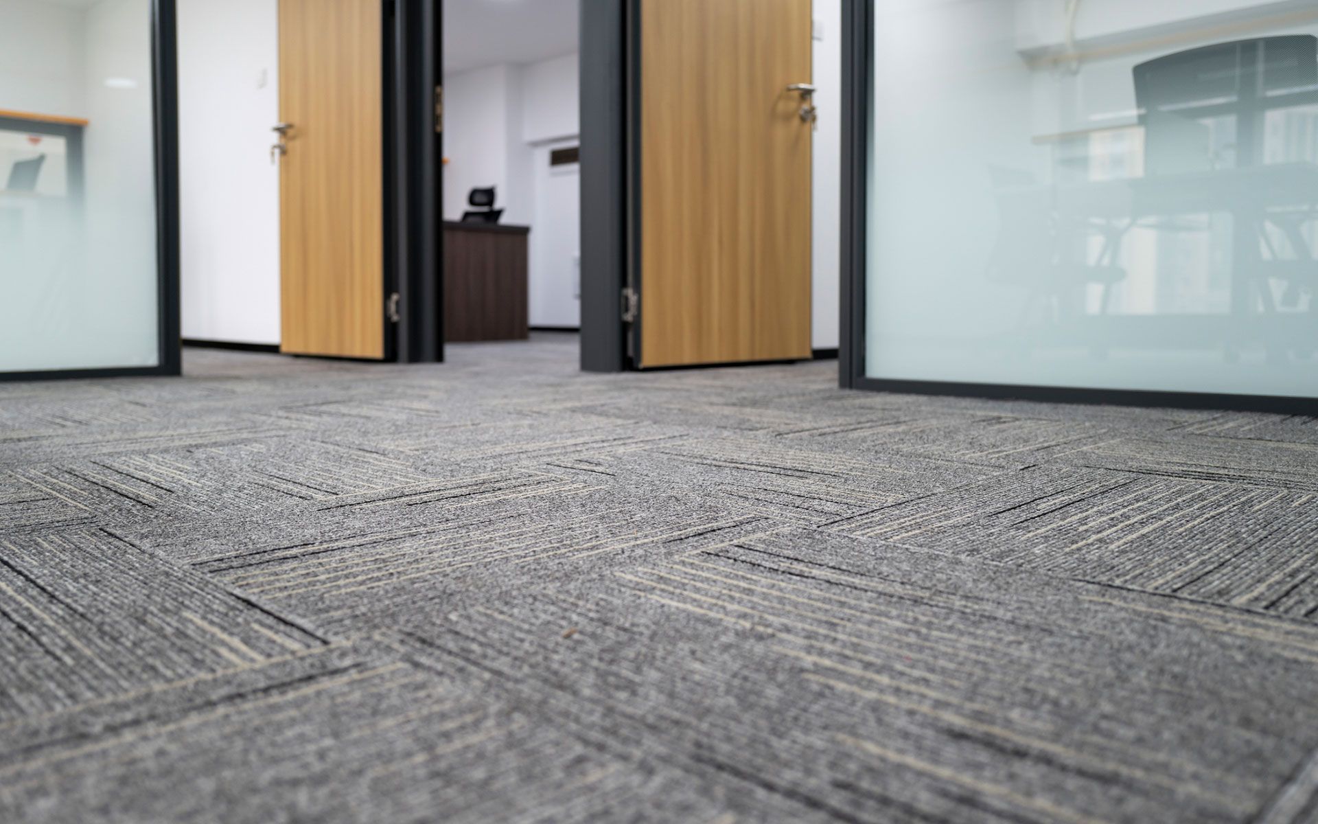 Person vacuuming carpeted office floor. Beige chairs, black pants, white shoes, and a glass wall visible.