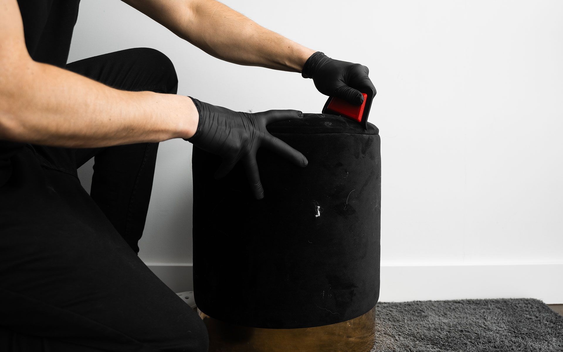 Person in black gloves placing a red tool inside a black cylindrical ottoman near a white wall.