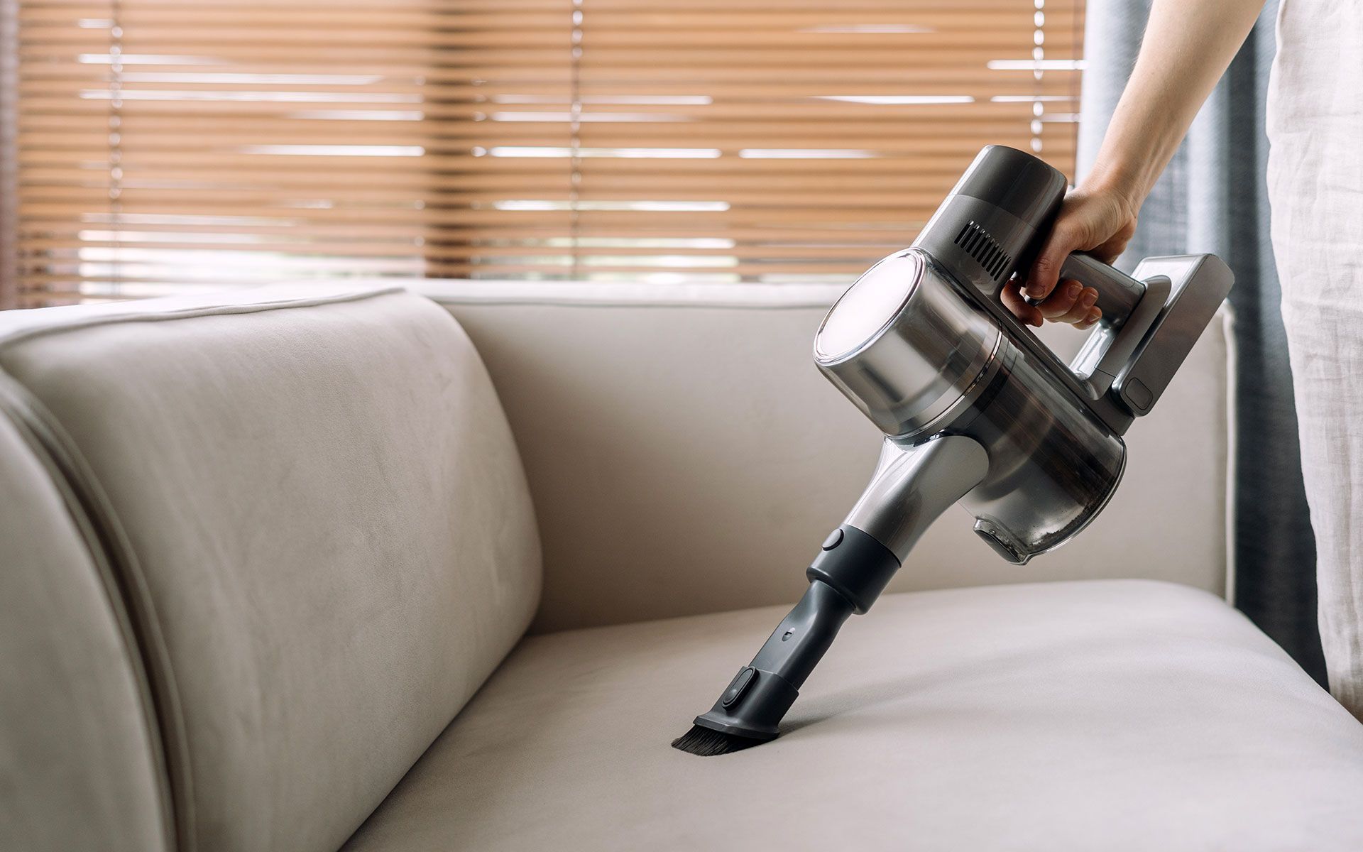 Person vacuuming a light-colored couch with a handheld vacuum, close-up.