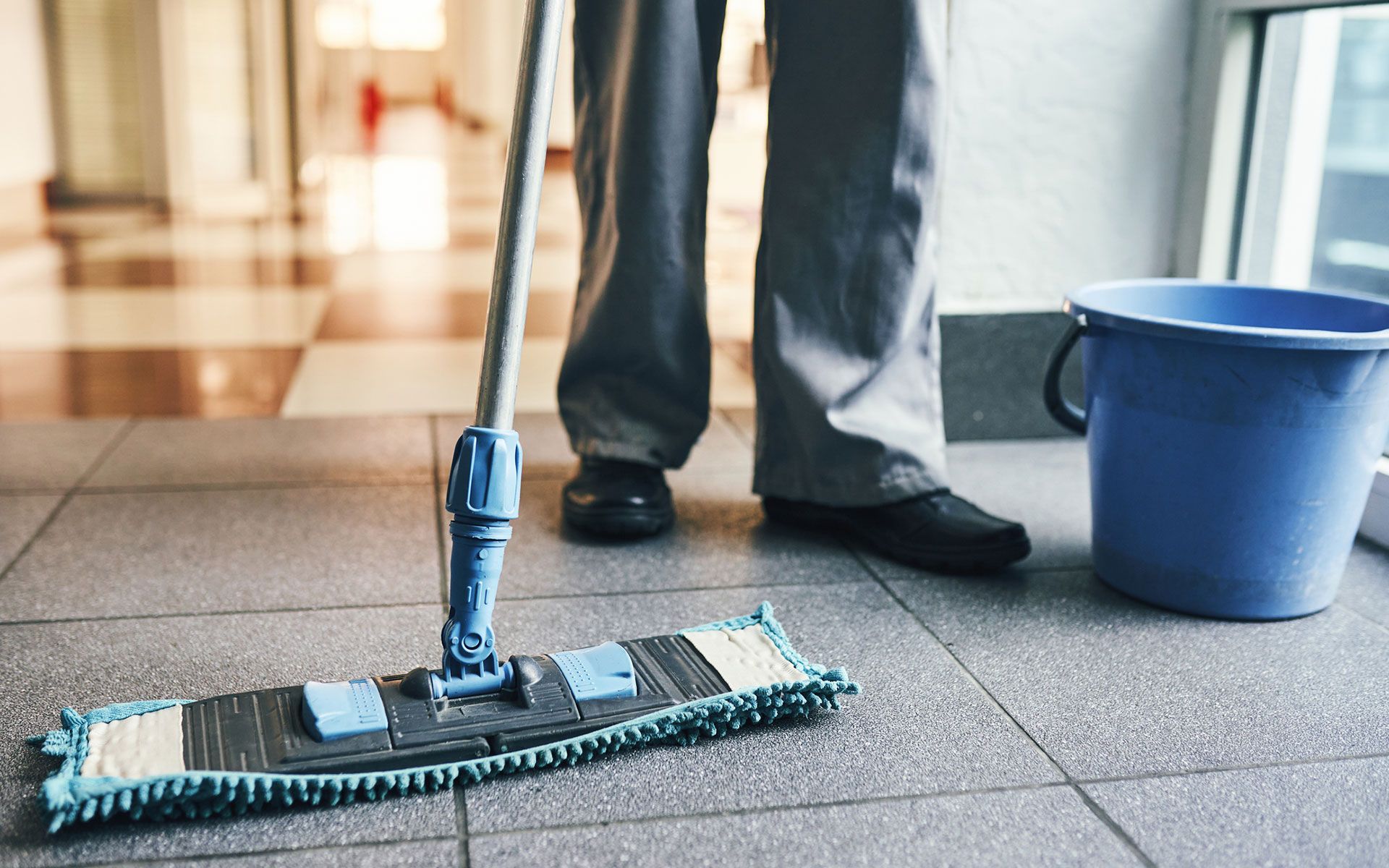 Person mopping a tiled floor next to a blue bucket, in a hallway.