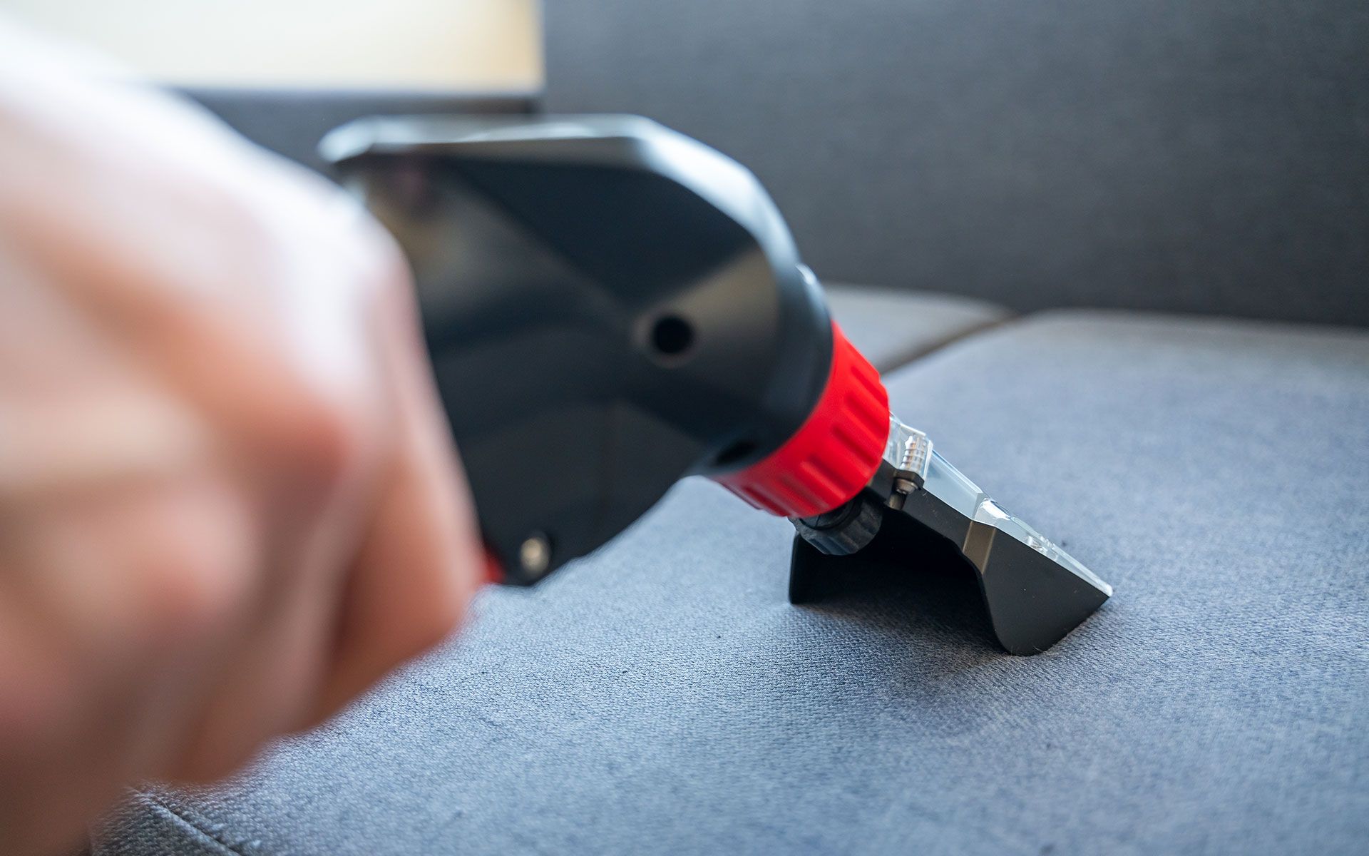 Person using a black and red upholstery cleaner on a dark grey couch.