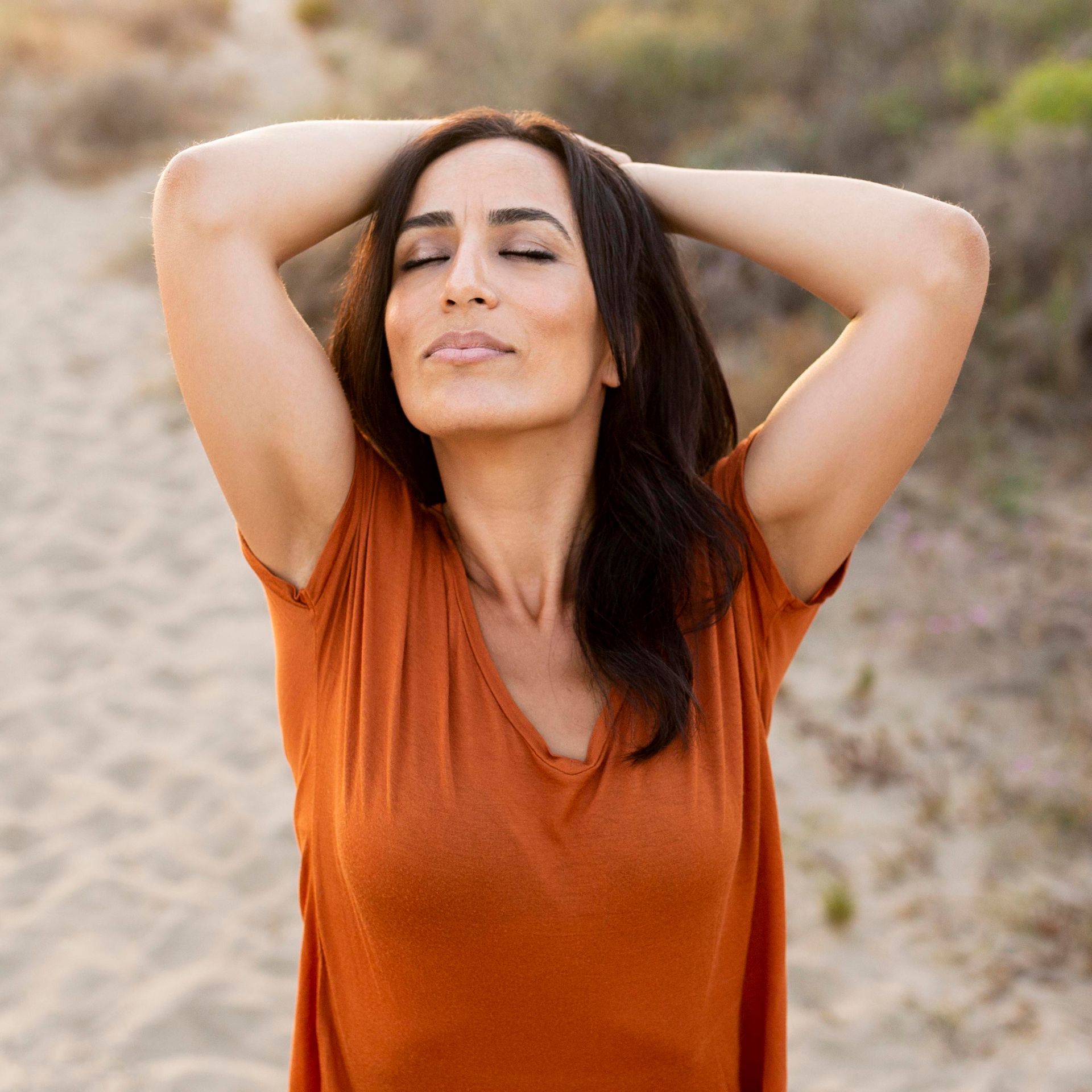 A woman in an orange shirt is standing on a beach with her eyes closed.