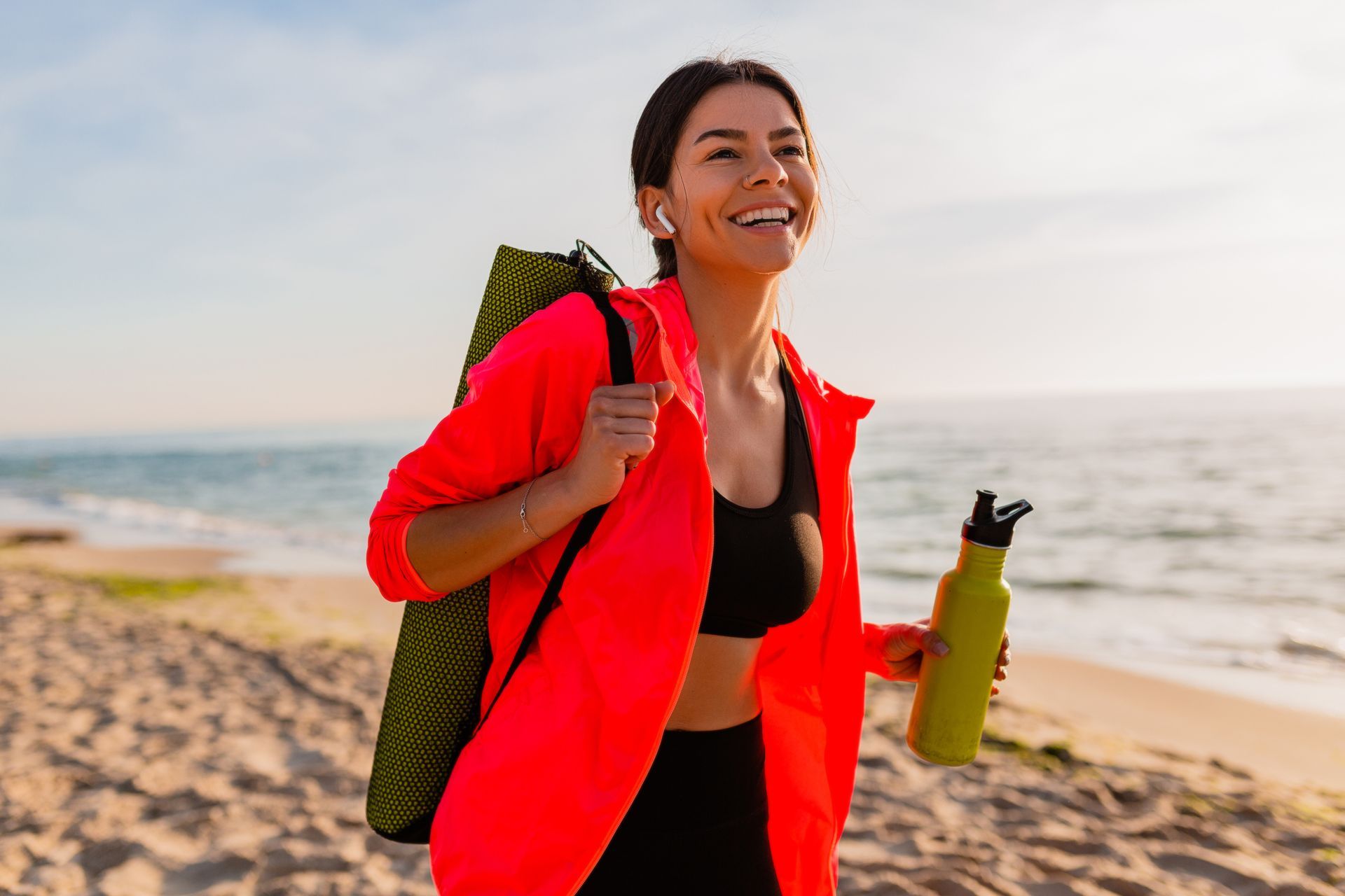 A woman is walking on the beach with a yoga mat and a water bottle.