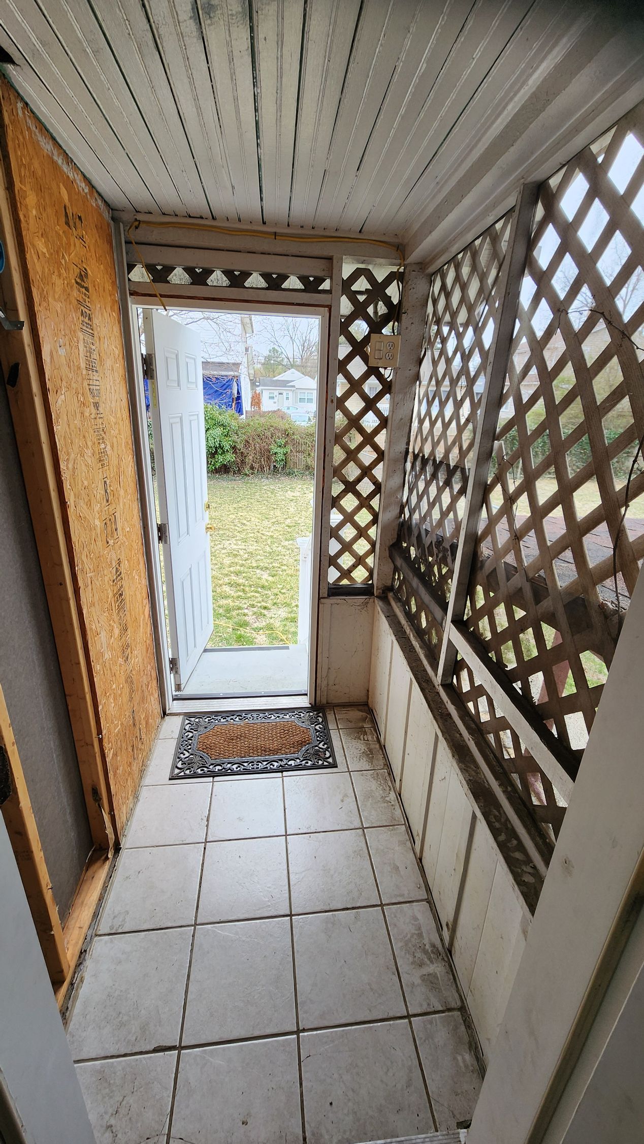 A porch with an open door and  a lattice work on the wall and tile floor.
