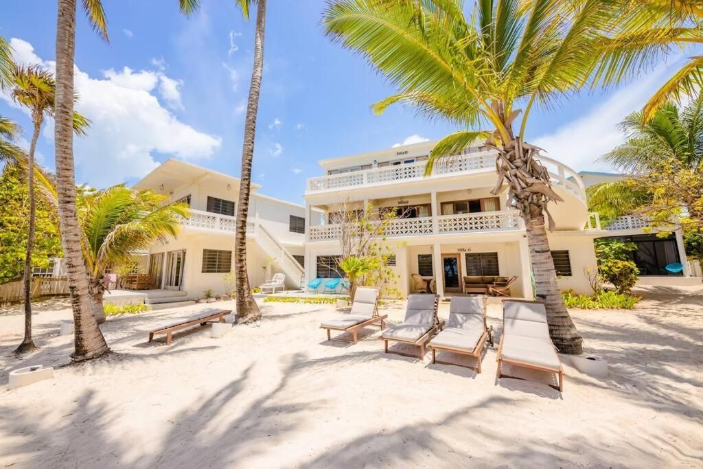 Beachfront resort with white buildings, palm trees, and lounge chairs under a blue sky.