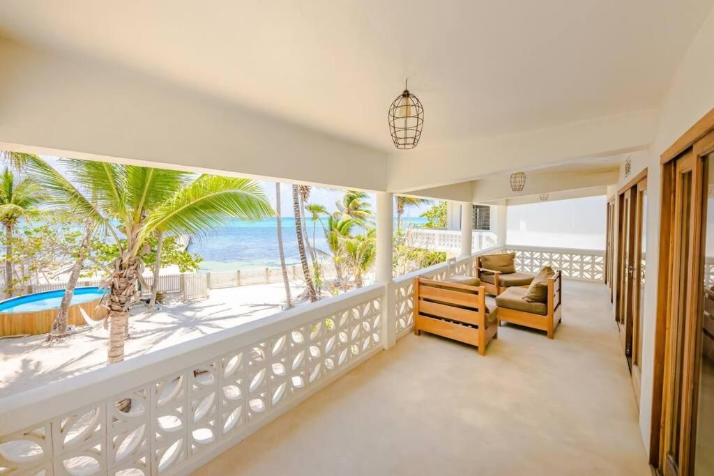 Balcony overlooking a beach with wooden furniture, palm trees, and a pool.