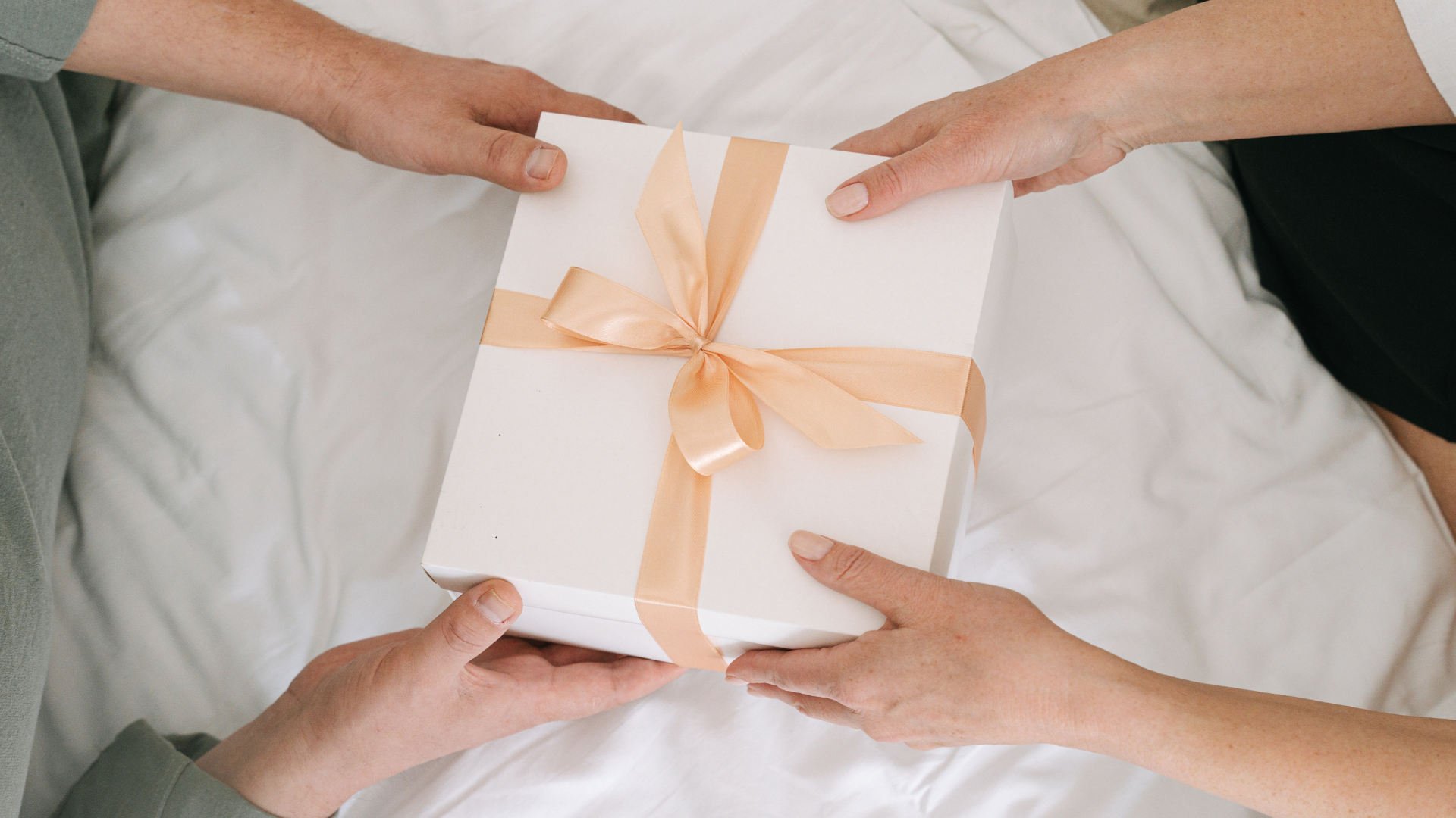 Two people exchanging a white gift box with a soft orange ribbon while sitting on a white bed.
