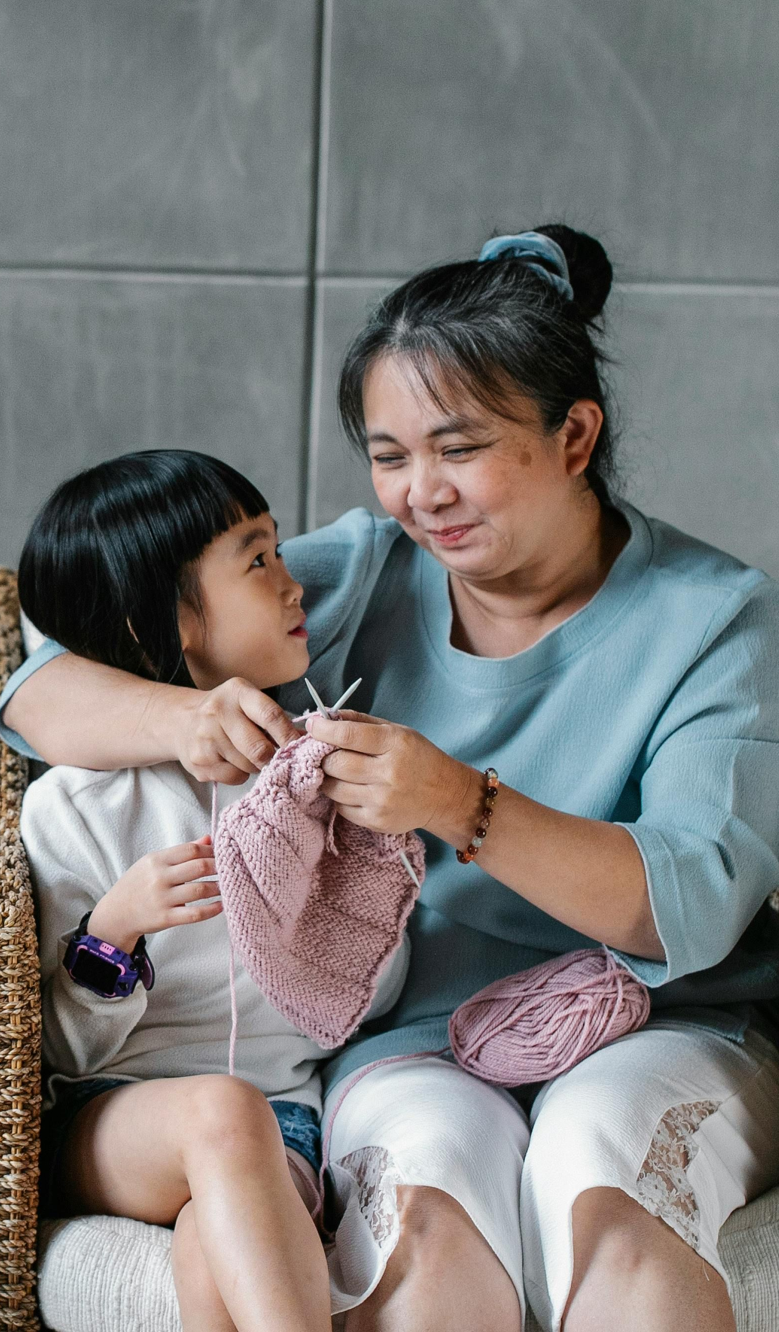 Woman teaching a child to knit on a wicker chair, holding needles and pink yarn, looking down.