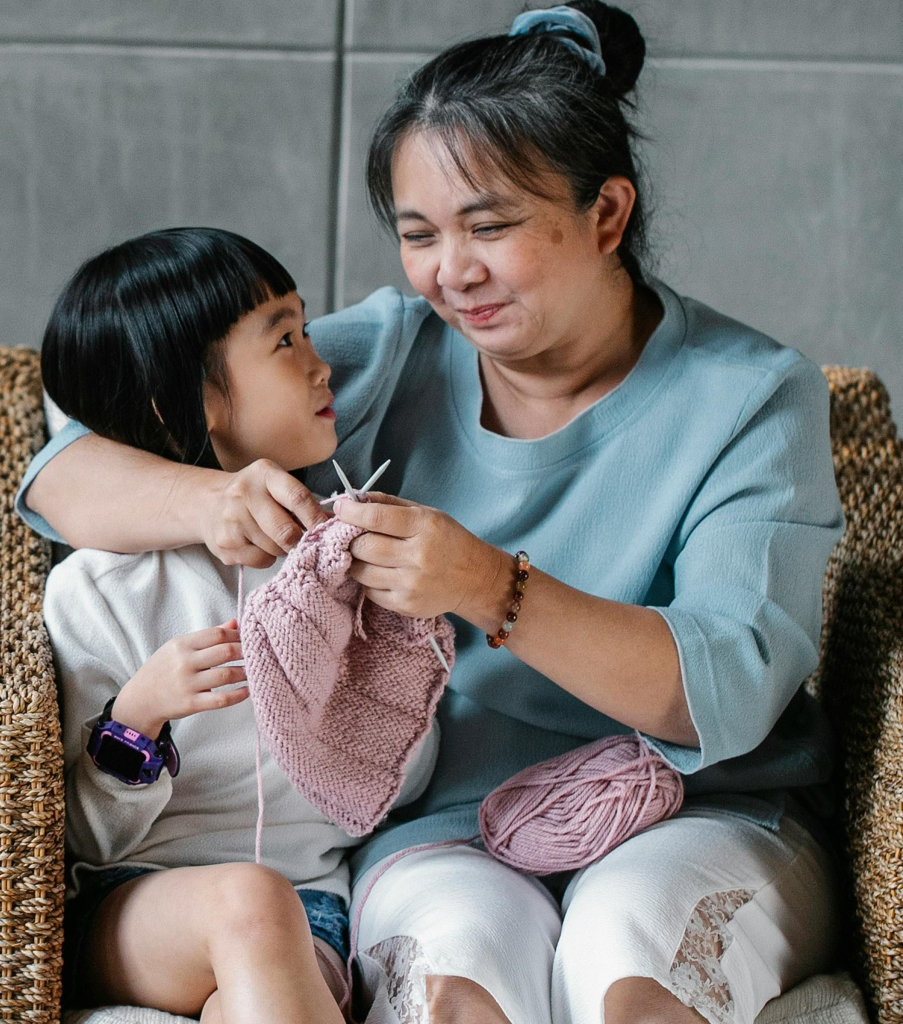 Woman teaching a child to knit on a wicker chair, holding needles and pink yarn, looking down.