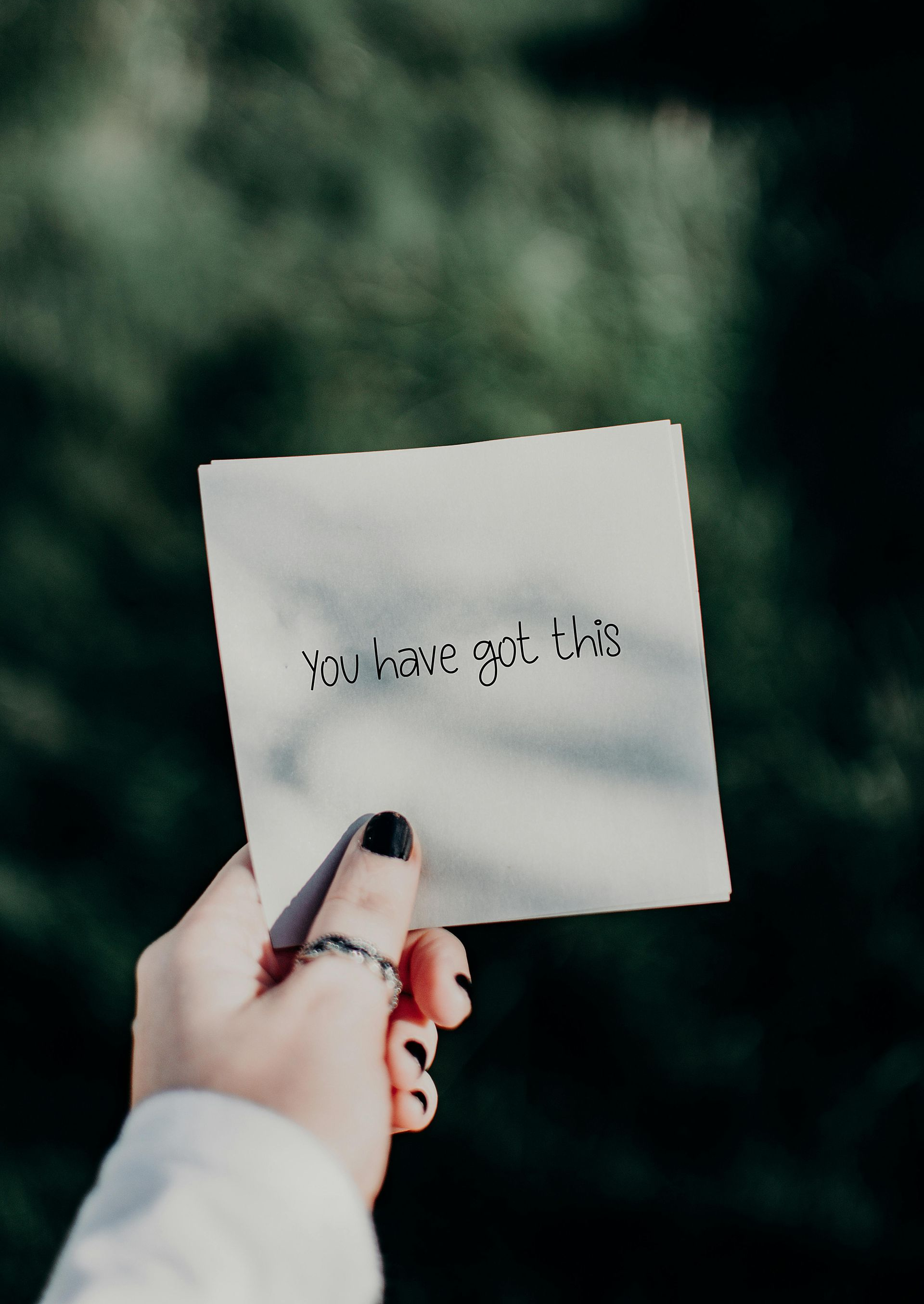 Person's hand holding a square card with text against a blurred green background. Black nail polish and ring visible.