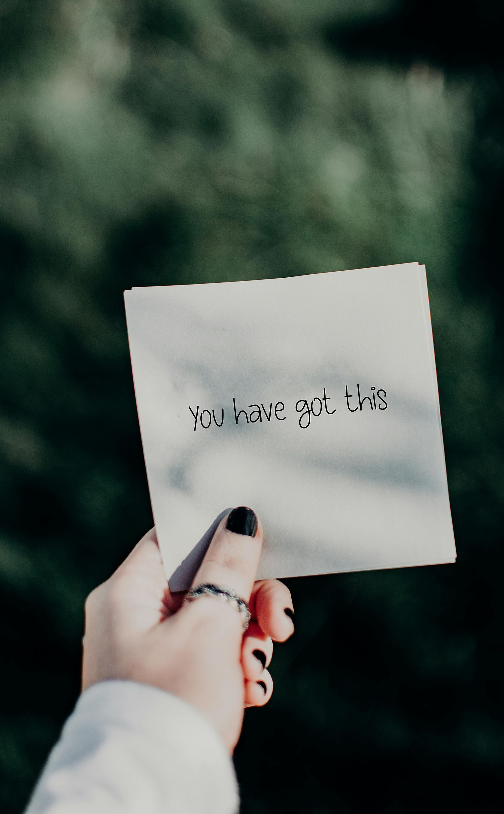 Person's hand holding a square card with text against a blurred green background. Black nail polish and ring visible.
