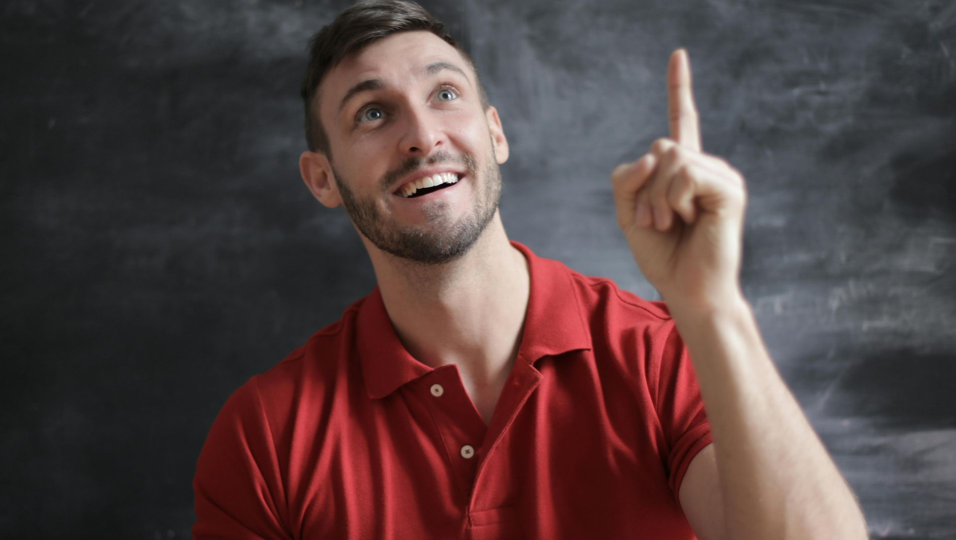 Man in red shirt points upward with an excited expression in front of a dark background.