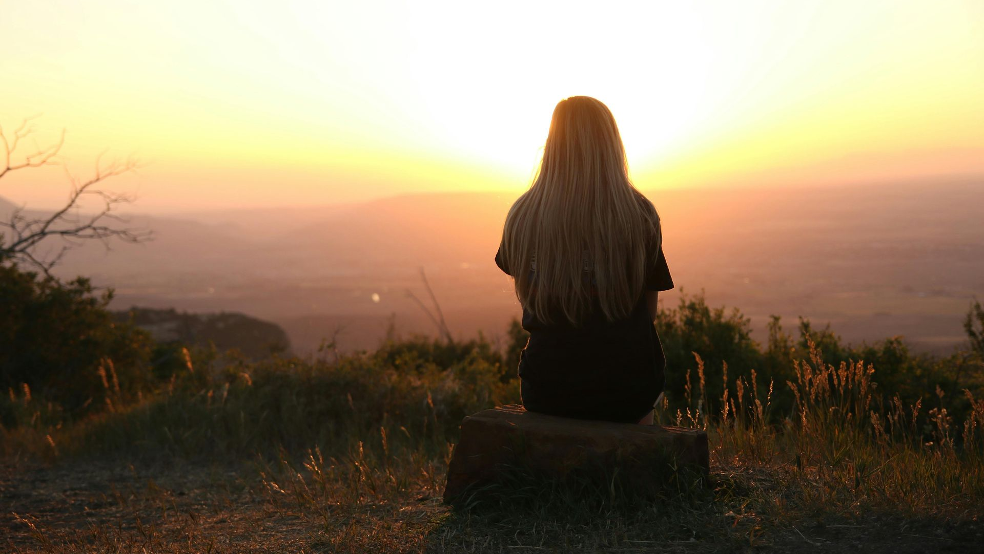Woman with long blonde hair sits atop a hill, back to camera, watching a sunset.