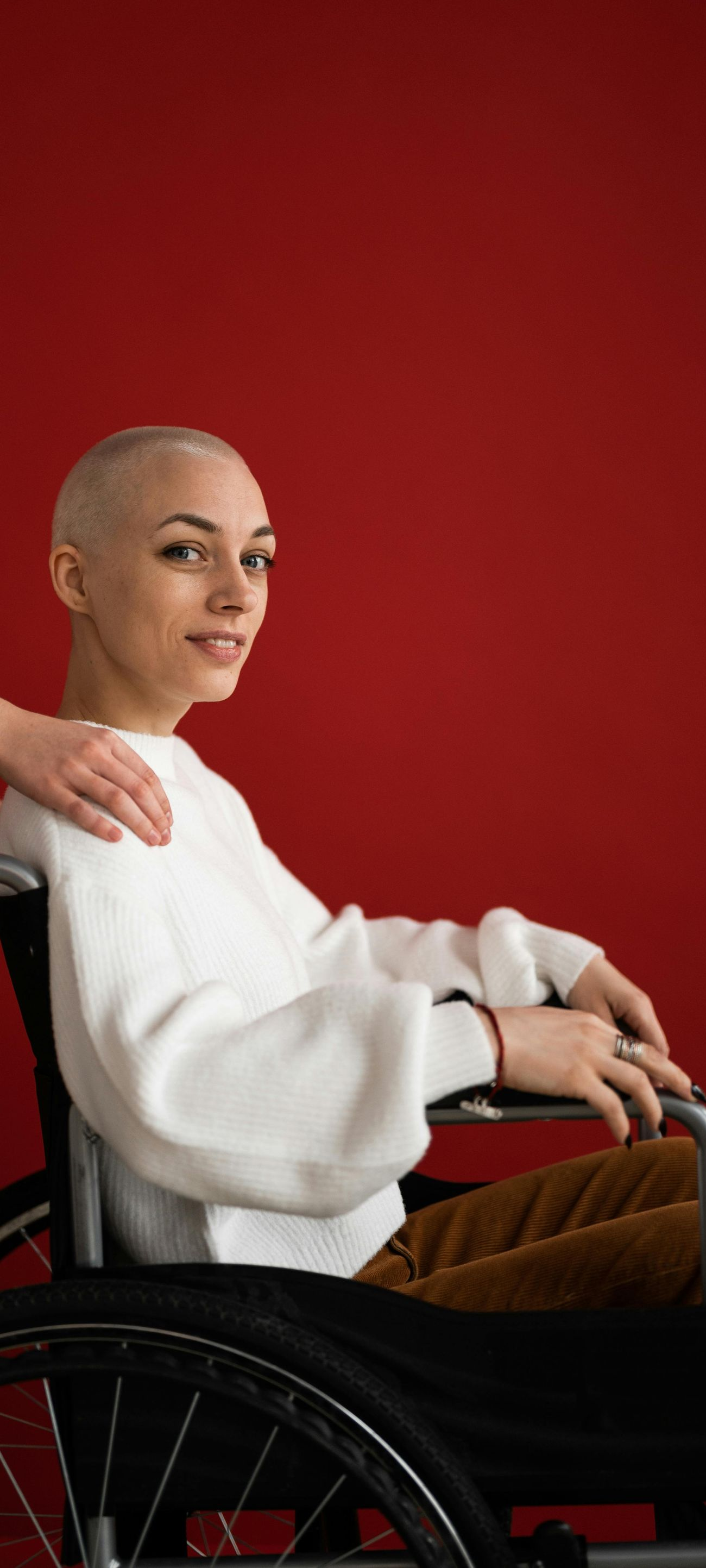 Woman in wheelchair with shaved head, hand on shoulder, looking towards the right, against a red background.