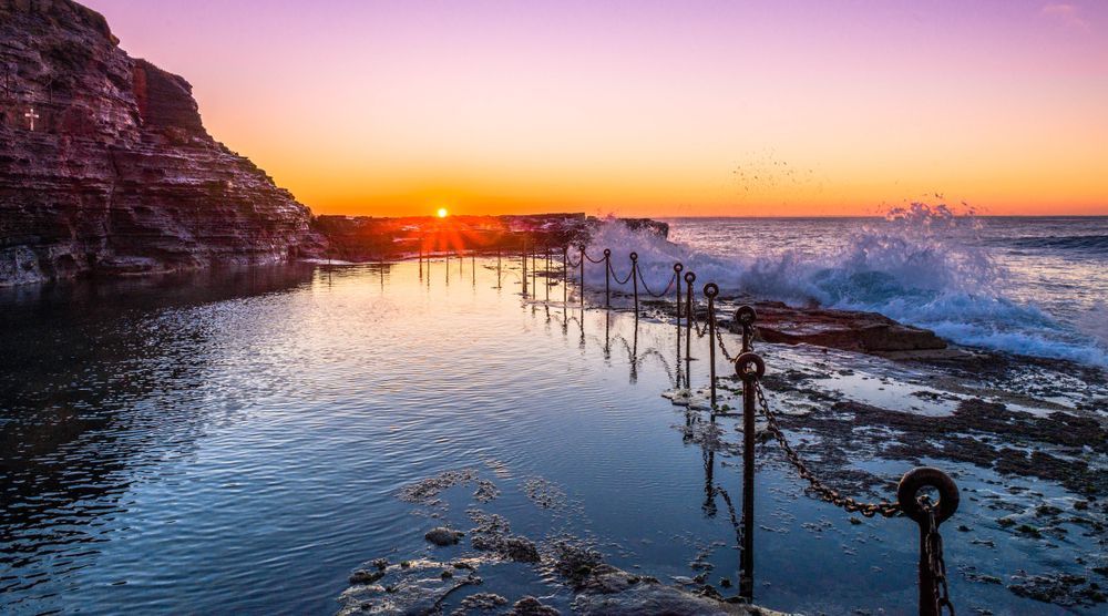A Sunset Over A Body Of Water With Waves Crashing On The Shore — CJ Electrical & Solar In Lambton, NSW
