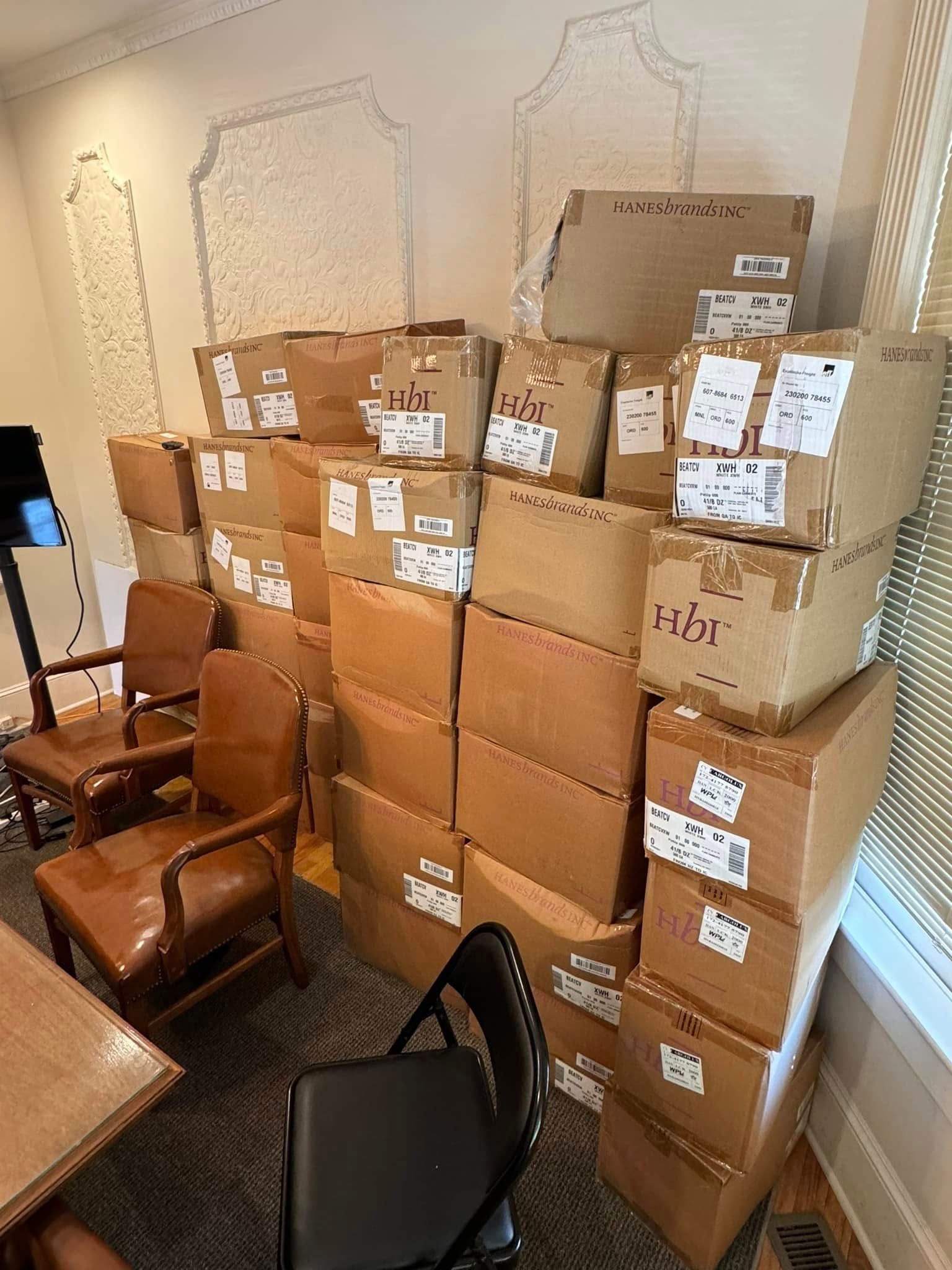 Pile of cardboard boxes in a room with two chairs, near a window with blinds.