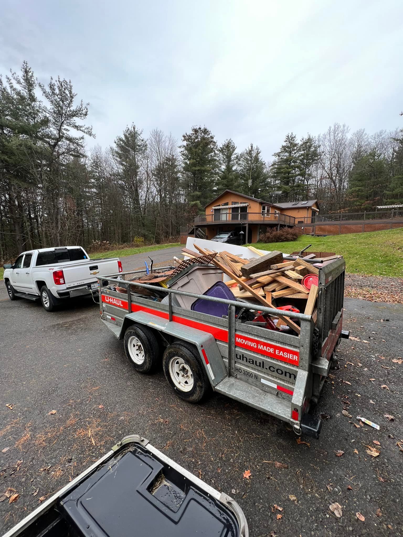 Truck pulling a trailer loaded with wood debris. A house and trees are in the background on a cloudy day.