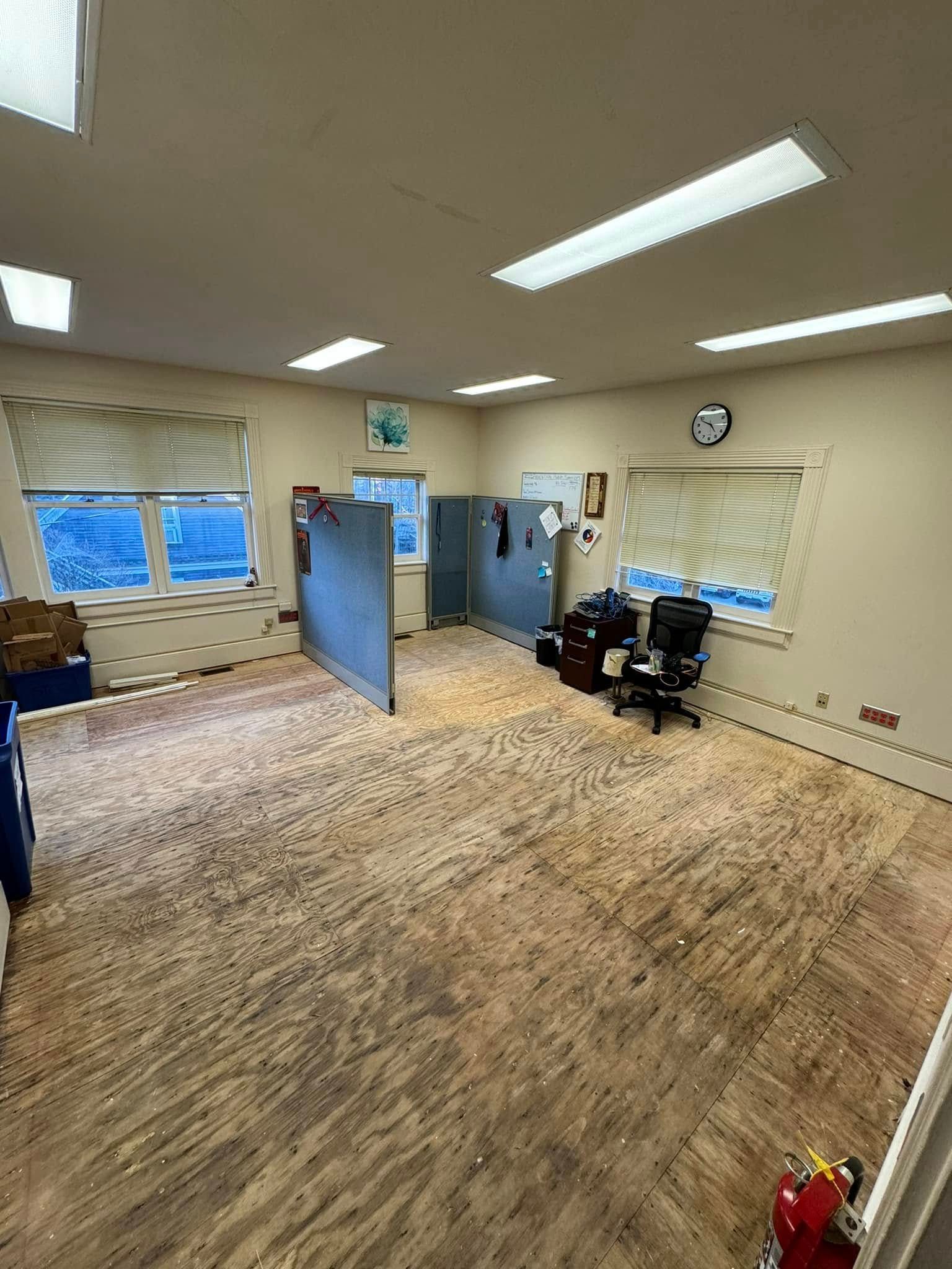 Empty office with exposed plywood floor, two windows, two partition walls, and a fire extinguisher.