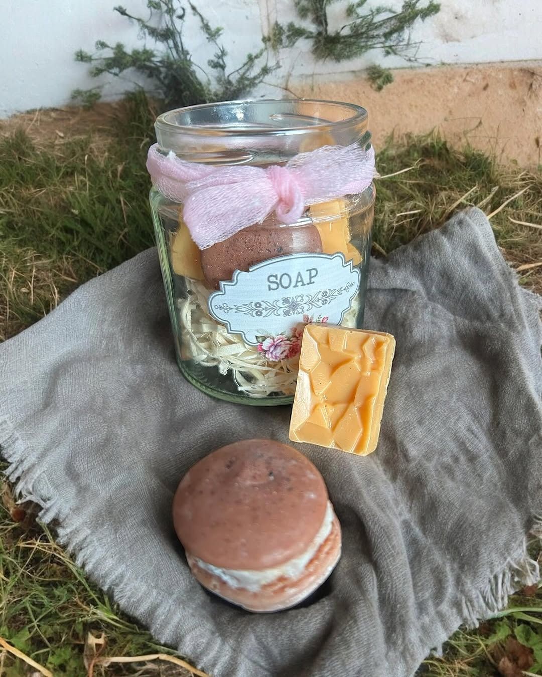 Tarro de jabones artesanales con cinta, sobre un paño de lino. Un jabón tiene forma de macarrón. Exterior, césped.