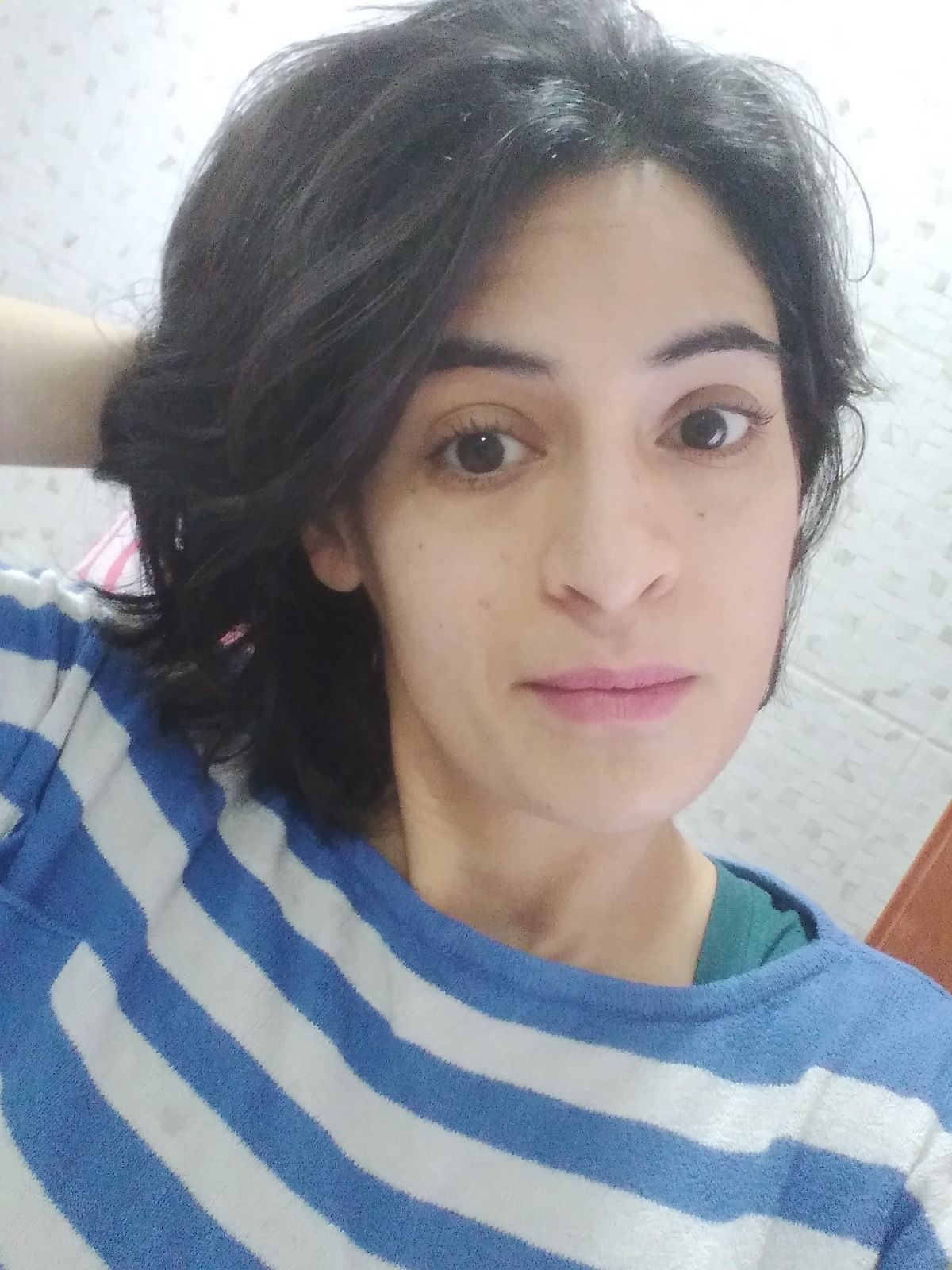 Mujer con cabello oscuro, camisa de rayas azules, mano en el cabello, mirando a la cámara con expresión neutra.