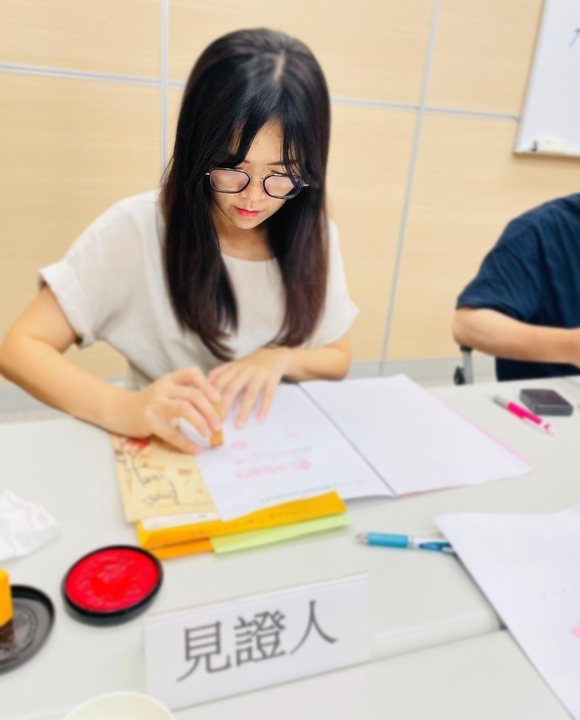 詠華地政士事務所所長-鄭秀慧女士在桌前蓋章。 「見證人」標誌清晰可見。桌上還有紅色印泥和筆記本。
