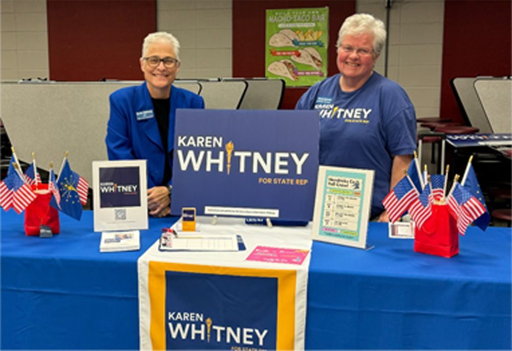 Two women at a political campaign table with signs and flags. Blue and white colors are prominent.