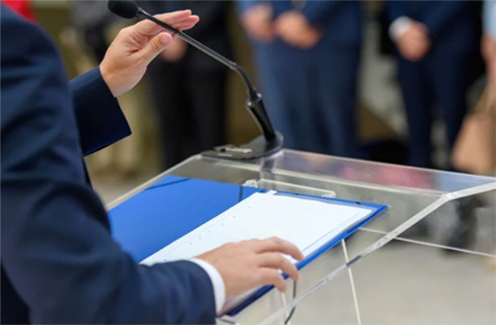 Person speaking at a podium with microphone, holding papers. Blue folder on the clear podium, blurred background.