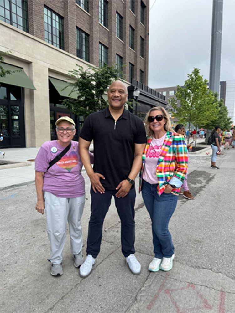 Three people standing on a city street. Woman in white pants, man in a black polo shirt, and woman in jeans pose.