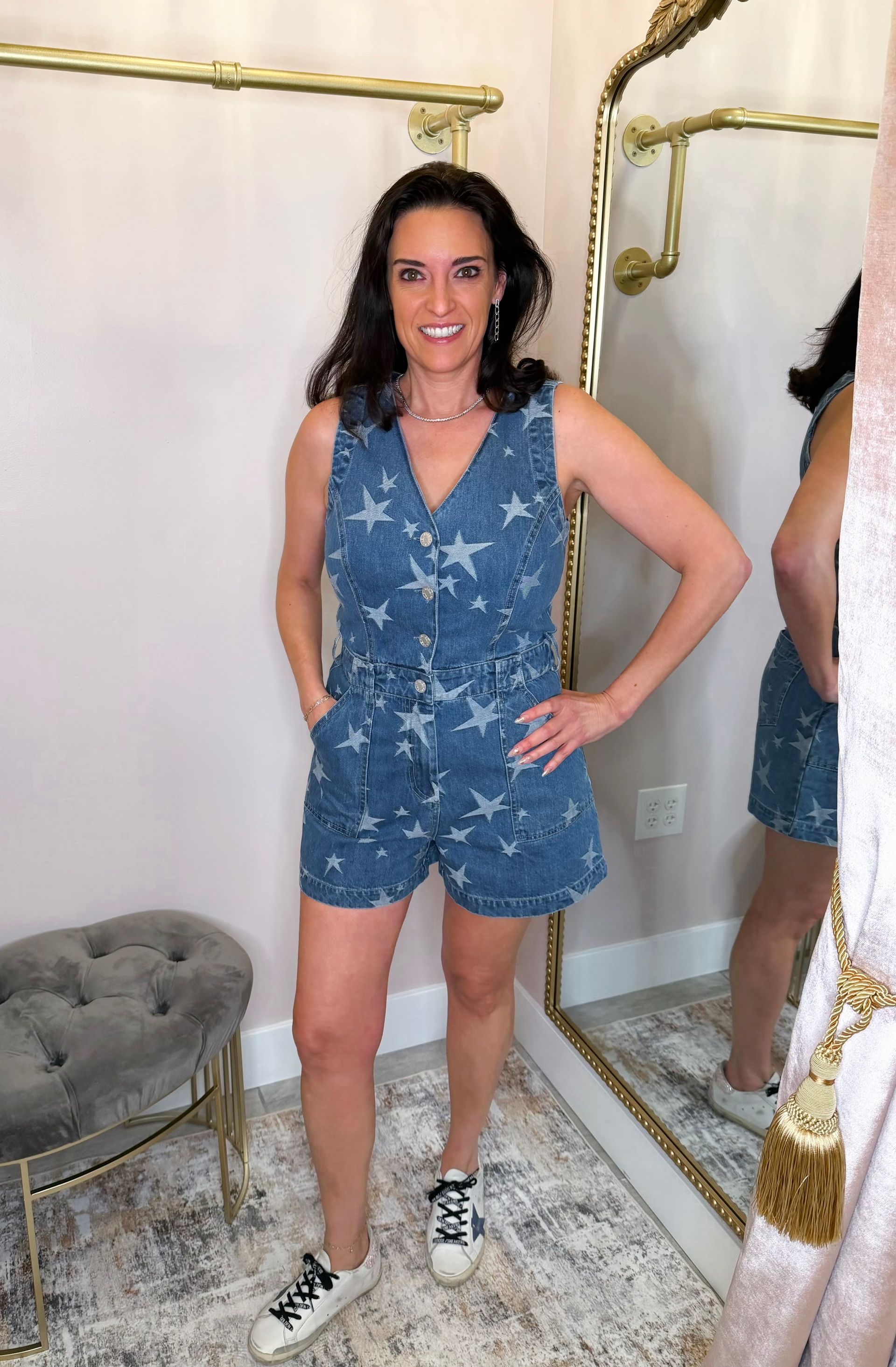 Person standing in a mirror-clad room, wearing a blue star-patterned romper and sneakers, smiling.