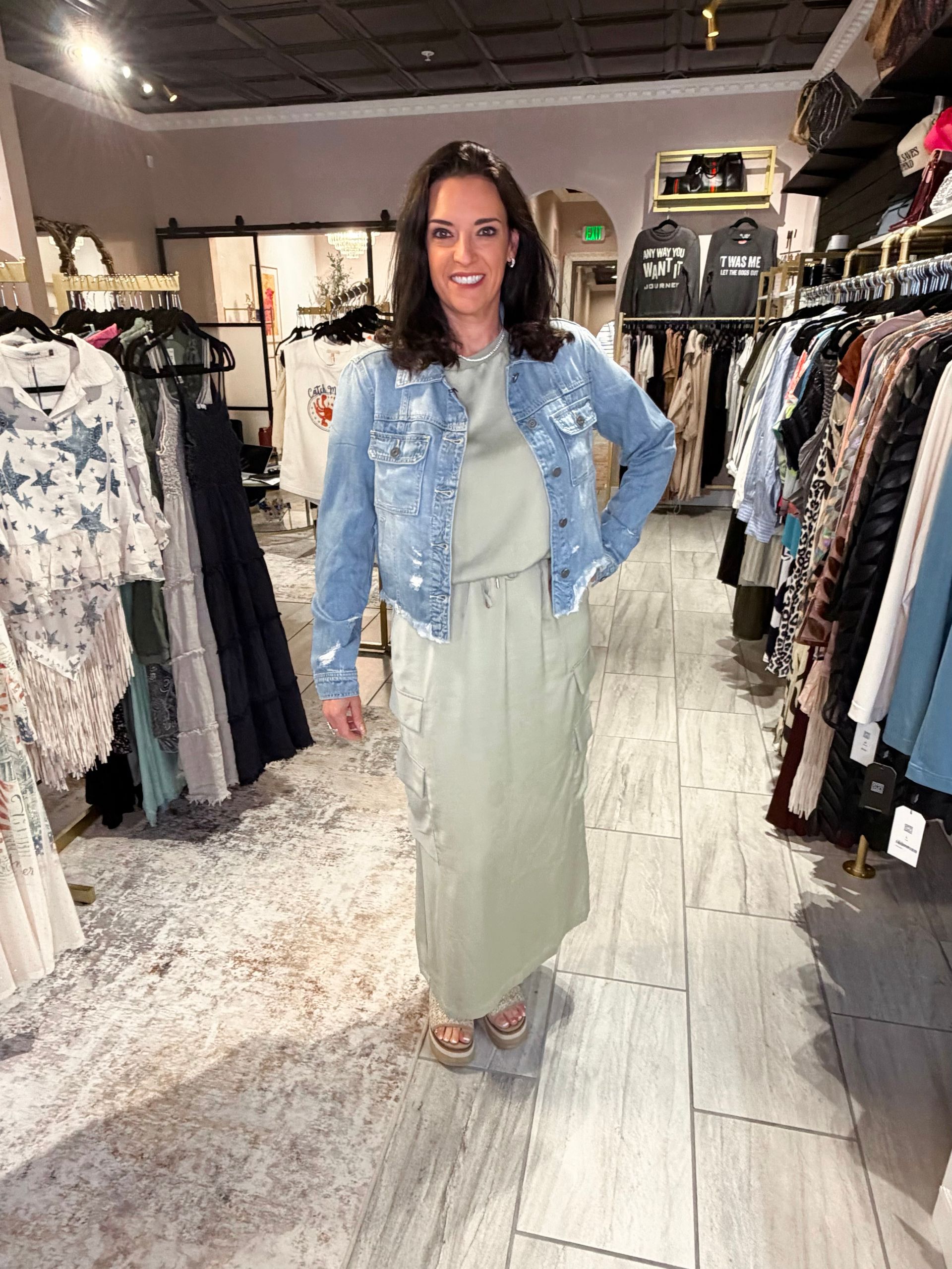 Woman standing in a clothing store, wearing a light blue denim jacket and long sage skirt.