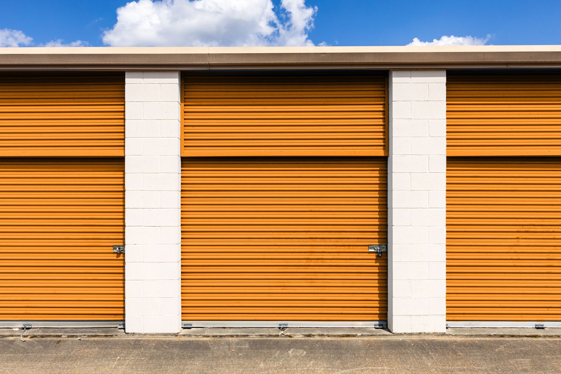 Exterior of outdoor self-storage units with orange roll-up doors and padlocks under blue sky. Exterior of outdoor self-storage units with orange roll-up doors and padlocks under blue sky.