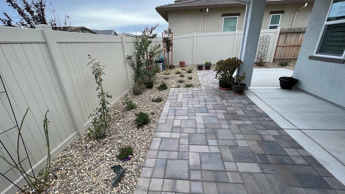 A brick walkway leading to a house with a white fence.