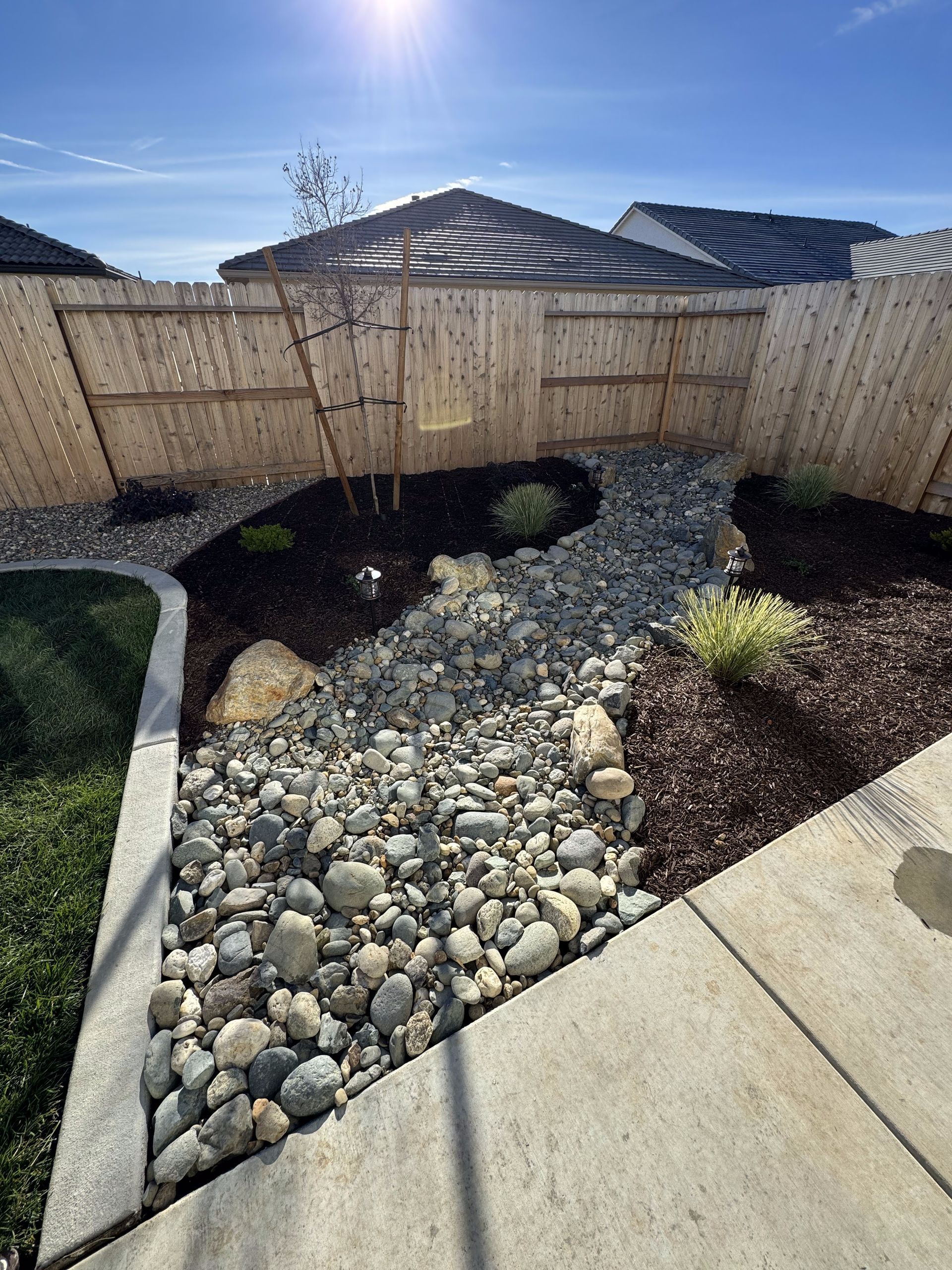 A backyard with a wooden fence , rocks , mulch and plants.