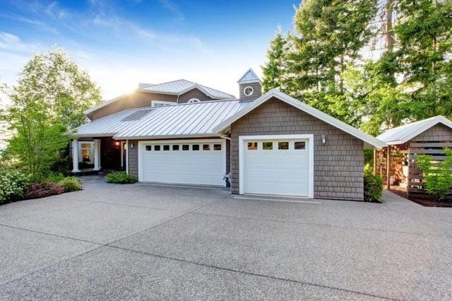 Gray house with white garage doors, asphalt driveway, and trees.