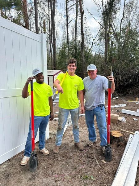 Three men are standing next to each other in front of a white fence holding shovels.