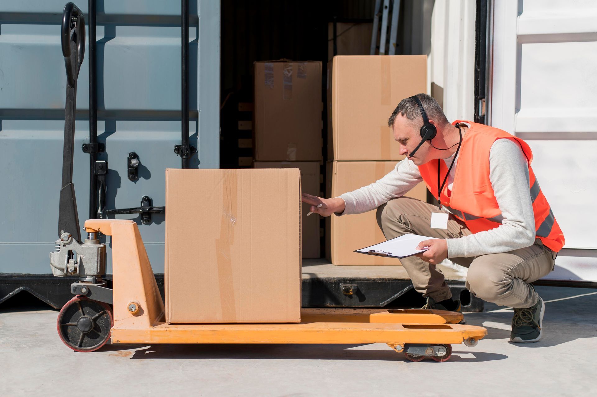 A man is kneeling down next to a pallet truck with boxes on it.