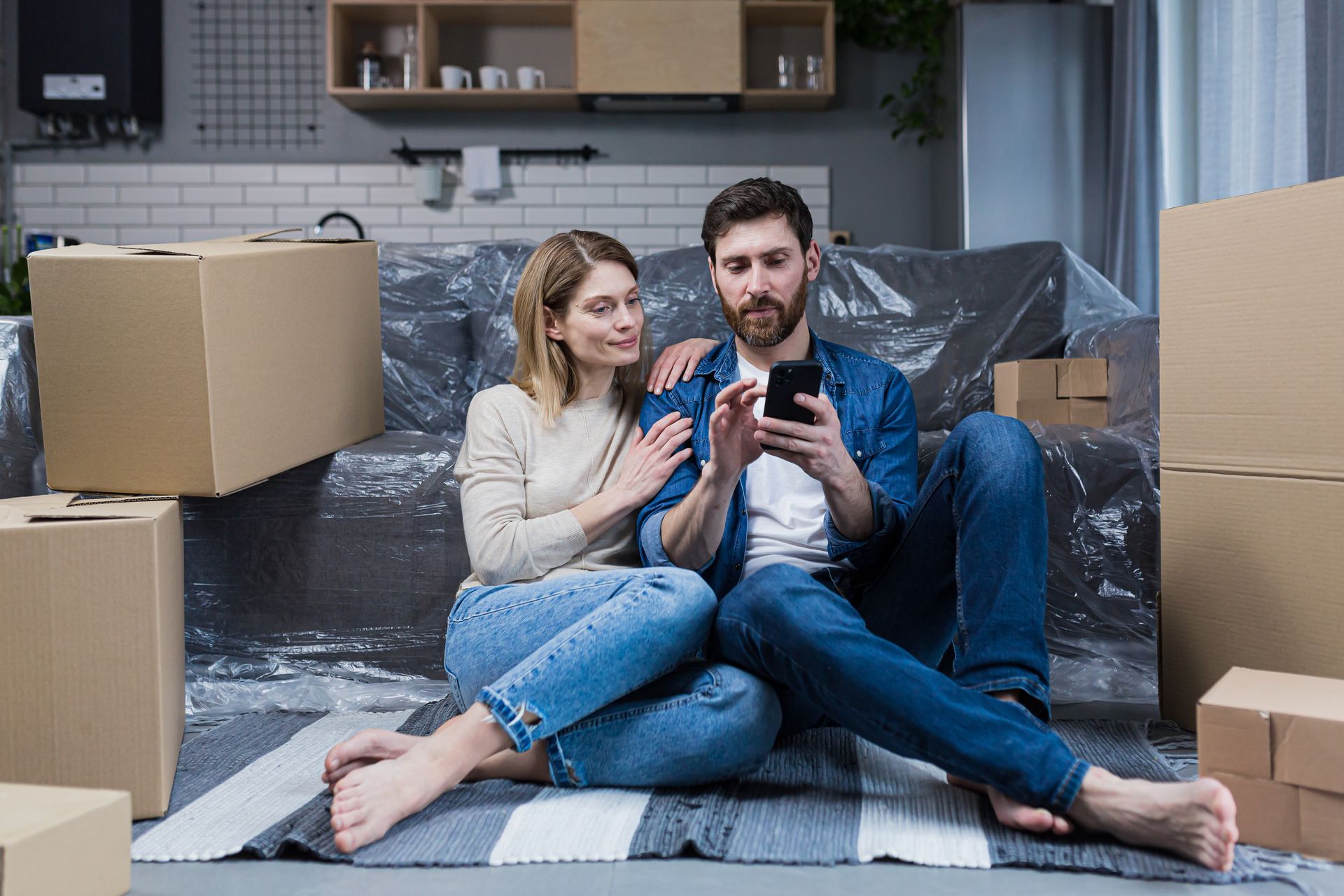 A man and a woman are sitting on the floor in a living room surrounded by cardboard boxes.