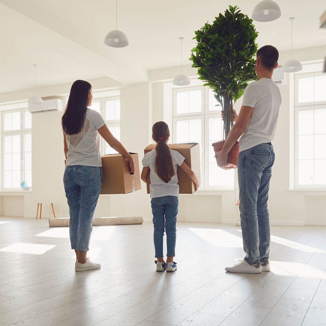 A family is standing in an empty room holding boxes and a potted plant.