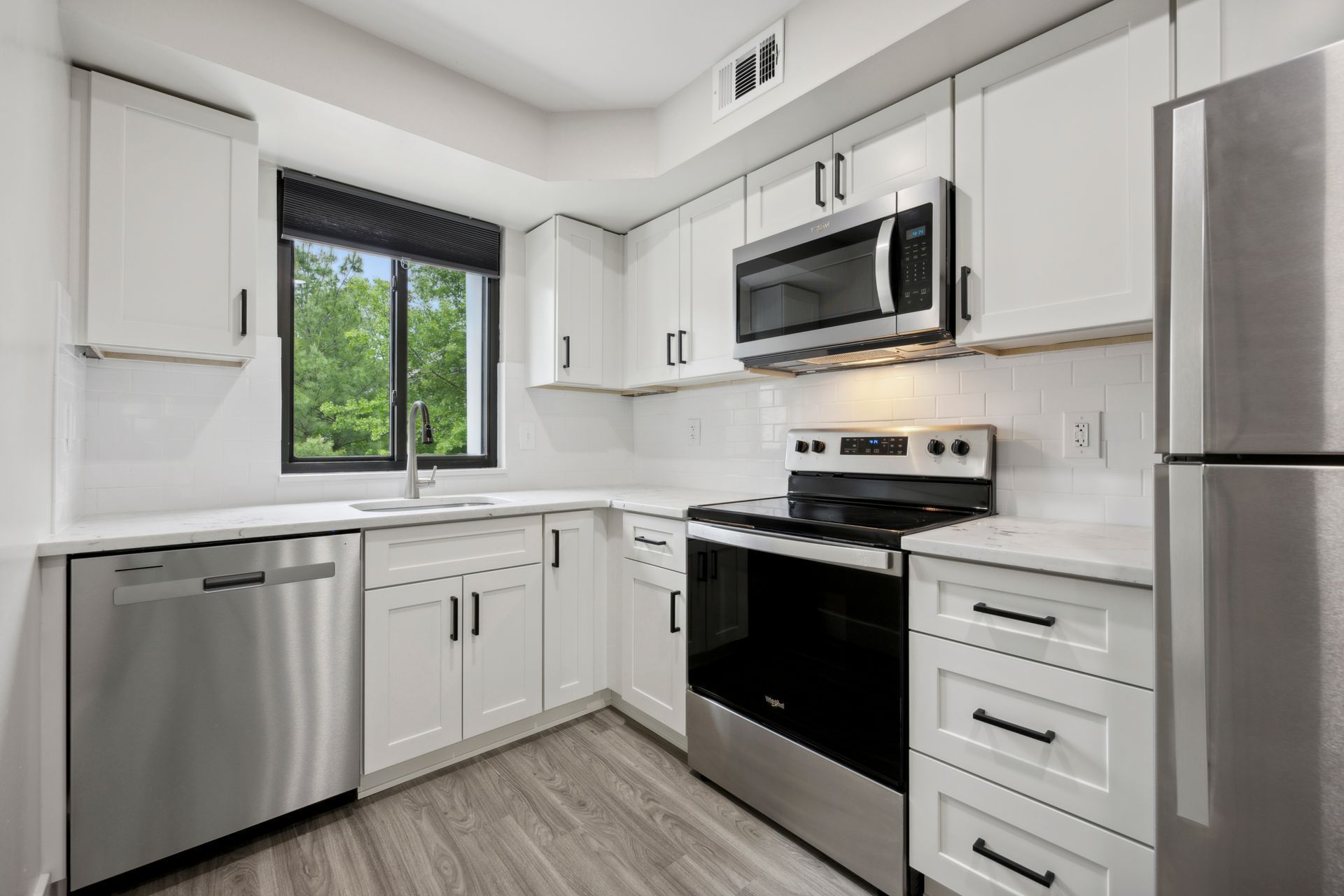 White kitchen with stainless steel appliances and cabinets, window overlooking greenery.