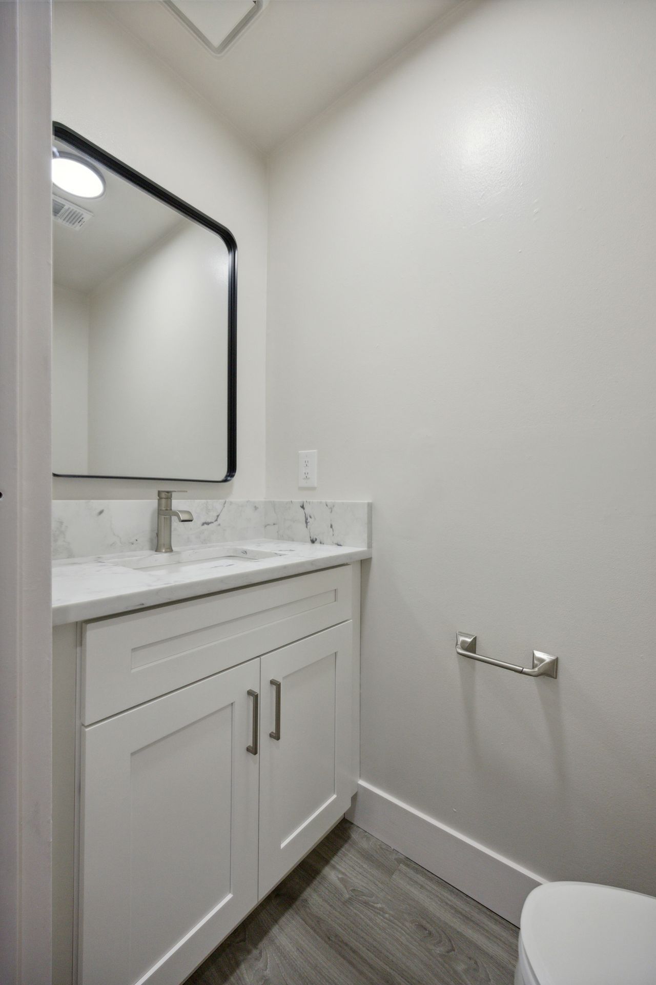 White bathroom with vanity, mirror, and toilet. Gray flooring, chrome towel rack, and white walls.