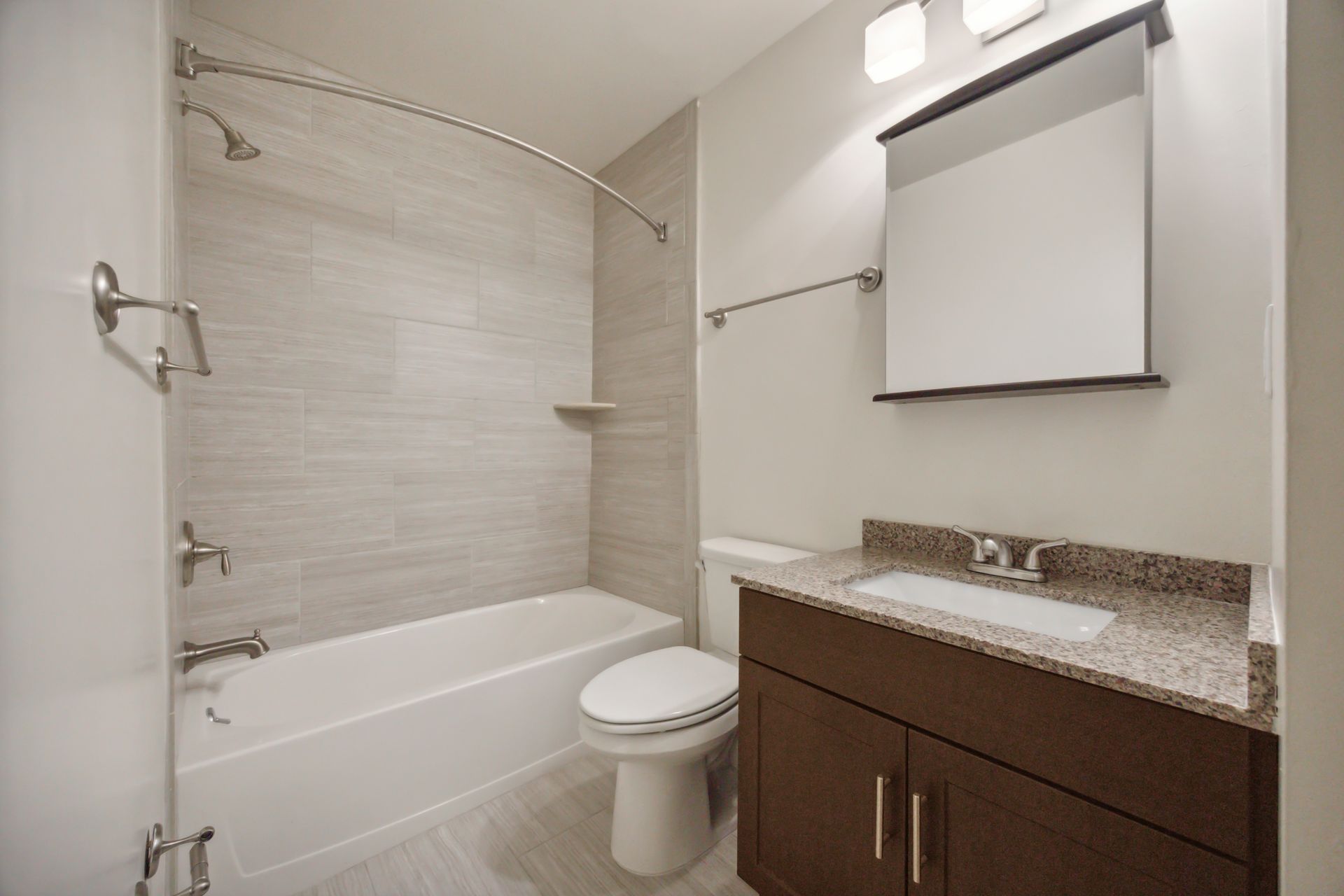 Bathroom with a white bathtub, toilet, vanity with granite countertop, and gray tiled walls.